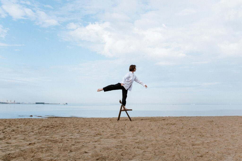 A male acrobat balances on a chair by the beach under a blue sky.