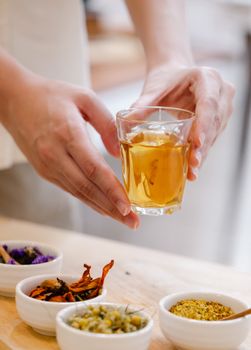 Close-up of hands holding herbal tea glass, surrounded by dried spices, in a cozy setting.